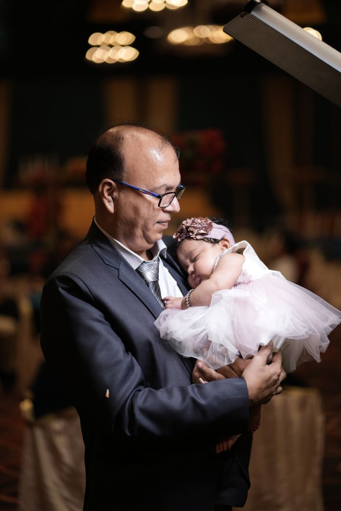 A man in a suit gently holds a sleeping baby in a festive indoor setting.