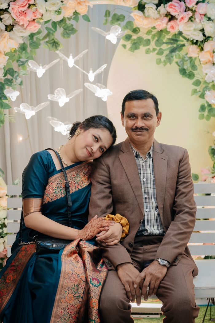 A couple in traditional attire posing affectionately under a floral arch at an outdoor event.