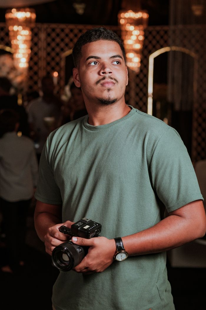 Confident young man holding camera, posing at an indoor event in warm lighting.