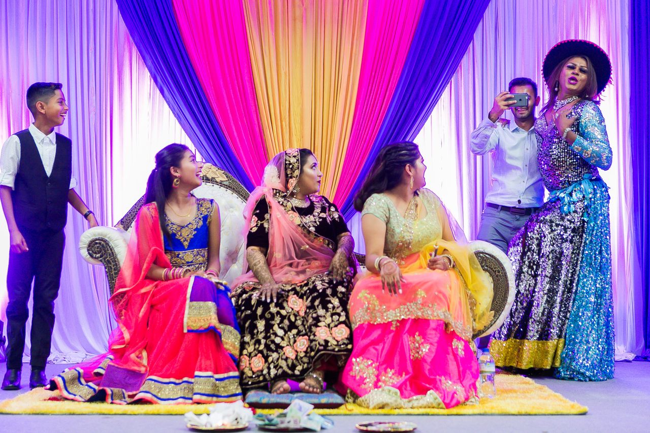 Full body of Indian women in traditional dresses sitting on sofa in room with colorful curtains and ethnic men during wedding ceremony